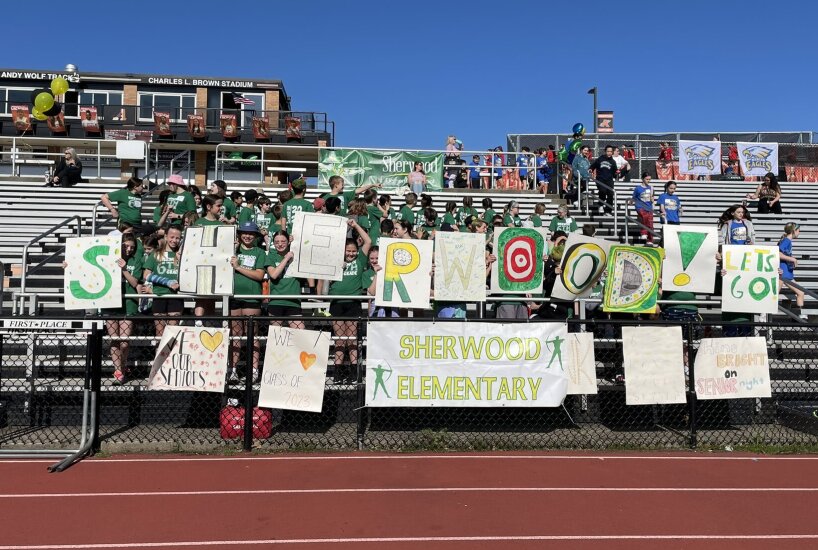 Students hold up signs that read Sherwood Elementary in the bleachers